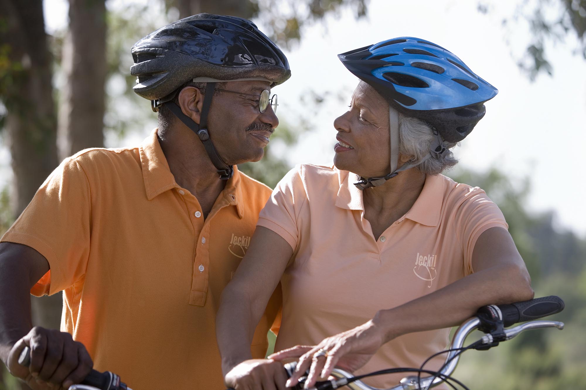Active senior couple wearing cycling helmets and polo shirts, sitting on bicycles in park, face to face, smiling